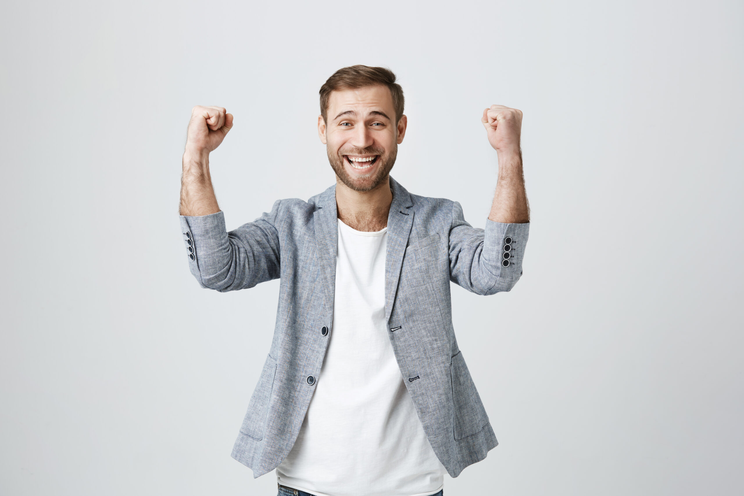 Happy lucky overjoyed male winner with stubble in trendy clothes clenches fists, rejoices triumph at work, glad to recieve high profits, has own prosperous business. Good-looking caucasian guy achieves goals